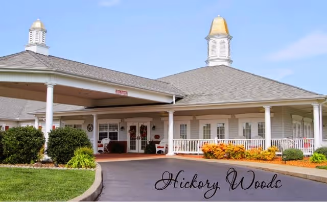 Exterior view of Hickory Woods Senior Living Community showing a single-story building with a covered entrance, white columns, and decorative cupolas on the roof. The landscaping includes bushes, green grass, and colorful flowers under a clear blue sky.