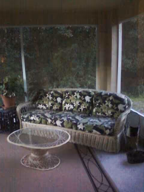 Wicker sofa with floral cushions and a glass round table on a sunlit screened porch.