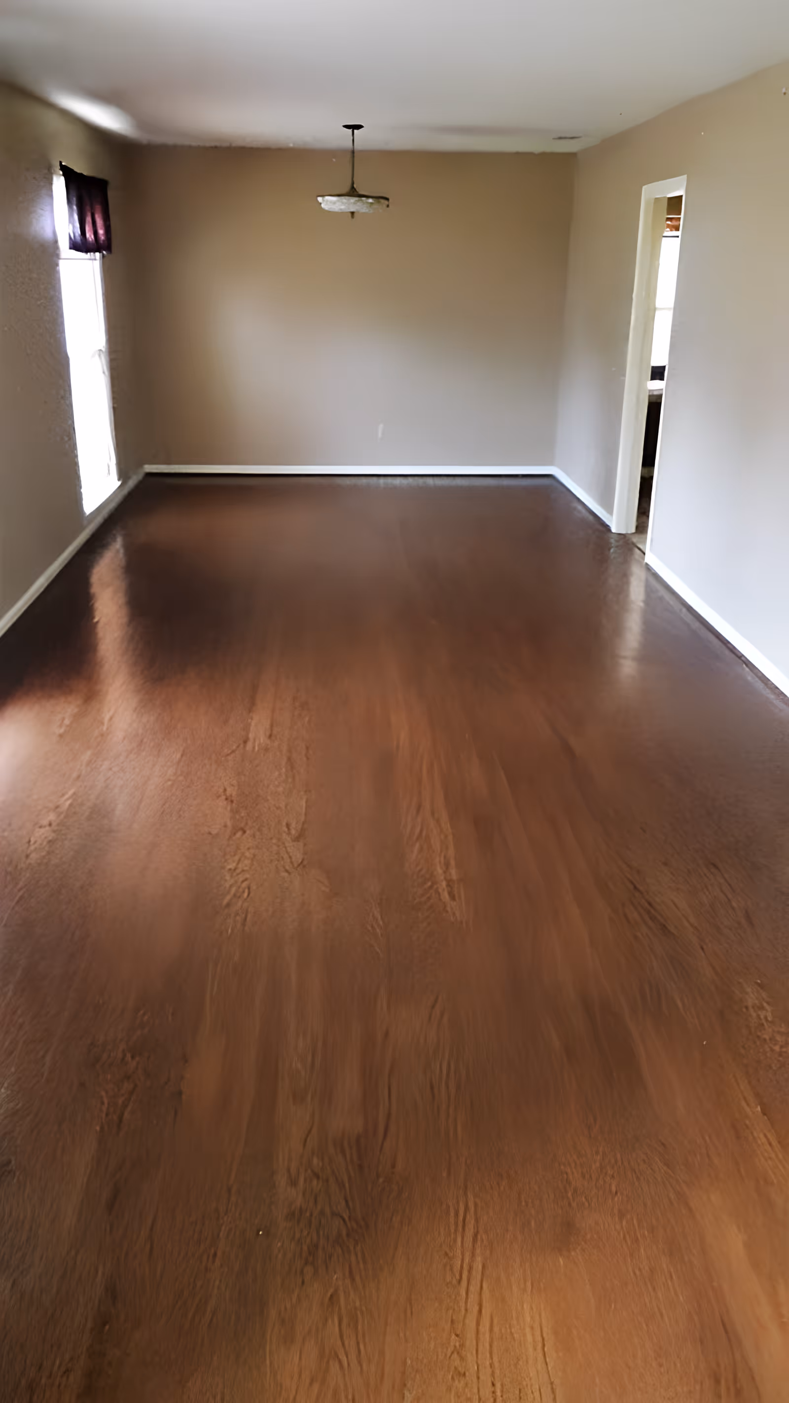 Empty room with polished wooden floor, beige walls, a single window with a dark curtain on the left, a ceiling light fixture, and an open doorway on the right leading to another room.