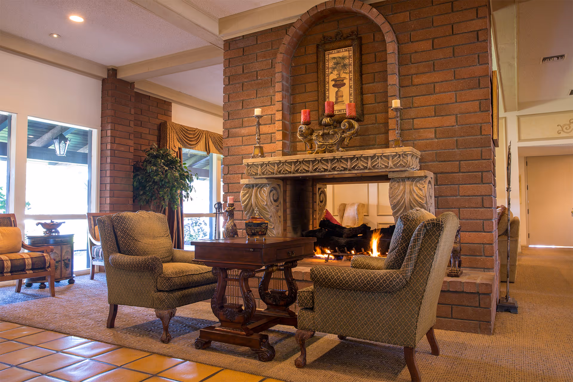 Cozy senior living community common area with a lit fireplace surrounded by a brick mantle, two upholstered armchairs facing each other with a wooden table in between, and additional seating and plants near large windows letting in natural light.
