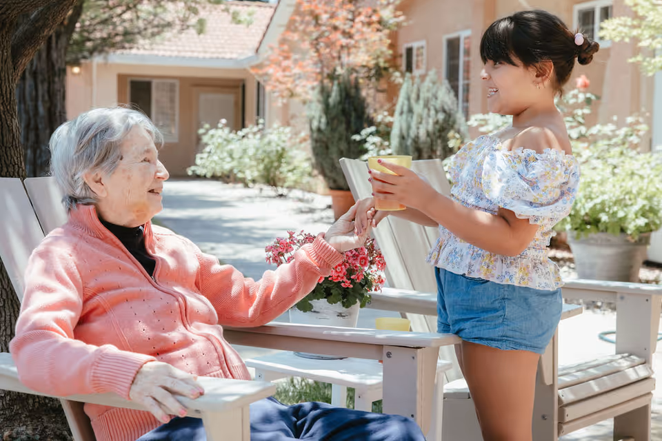 An elderly woman sitting in a wooden chair outdoors, smiling and holding hands with a young girl who is standing and handing her a yellow cup. They are in a garden area with plants and flowers, and a building with windows is visible in the background.
