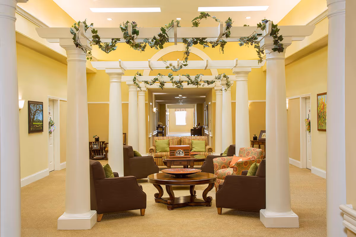 A cozy indoor seating area in a senior living facility with cream-colored columns and a white pergola adorned with green vines. The space features a round wooden coffee table surrounded by four brown armchairs with green pillows, and a striped sofa with green pillows in the background. The walls are painted yellow with white trim, and there are framed paintings and doorways along the hallway.