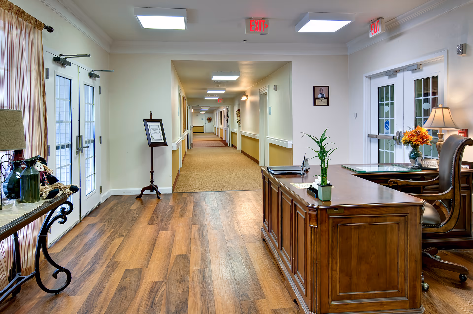 Reception area with a wooden desk, double entry doors, and a long carpeted hallway in a senior living facility.