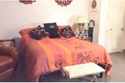 A bedroom with a large bed covered in an orange and red patterned bedspread, three decorative pillows, a small bench at the foot of the bed, a brown armchair to the left, a lamp on a nightstand, and a closed white door in the background.