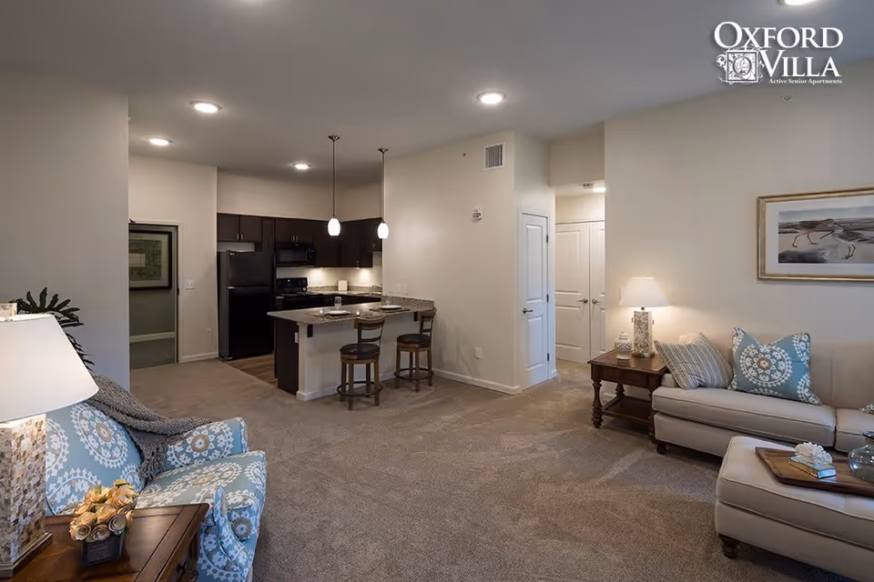 Interior view of a senior apartment living area with a beige carpet, a light beige sofa with patterned pillows, a matching ottoman, a wooden side table with a lamp, and a blue patterned armchair. The open kitchen features dark cabinets, a black refrigerator, a stove, and a granite countertop with two bar stools. The walls are light-colored, and there is a framed artwork on the wall above the sofa.