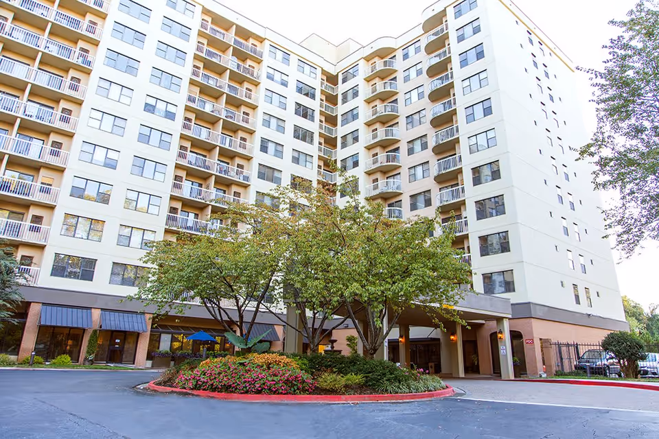 Exterior view of a multi-story retirement community building with balconies, a circular driveway, and landscaped greenery including trees and flowers in front of the entrance.