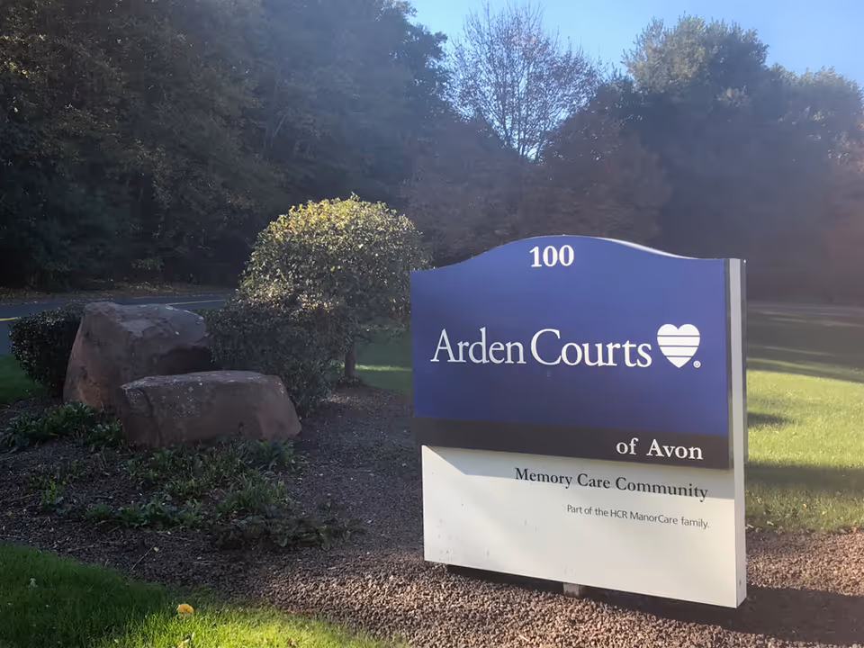 Outdoor view of a sign for Arden Courts of Avon Memory Care Community, located on a grassy area with bushes and large rocks nearby, surrounded by trees in the background.