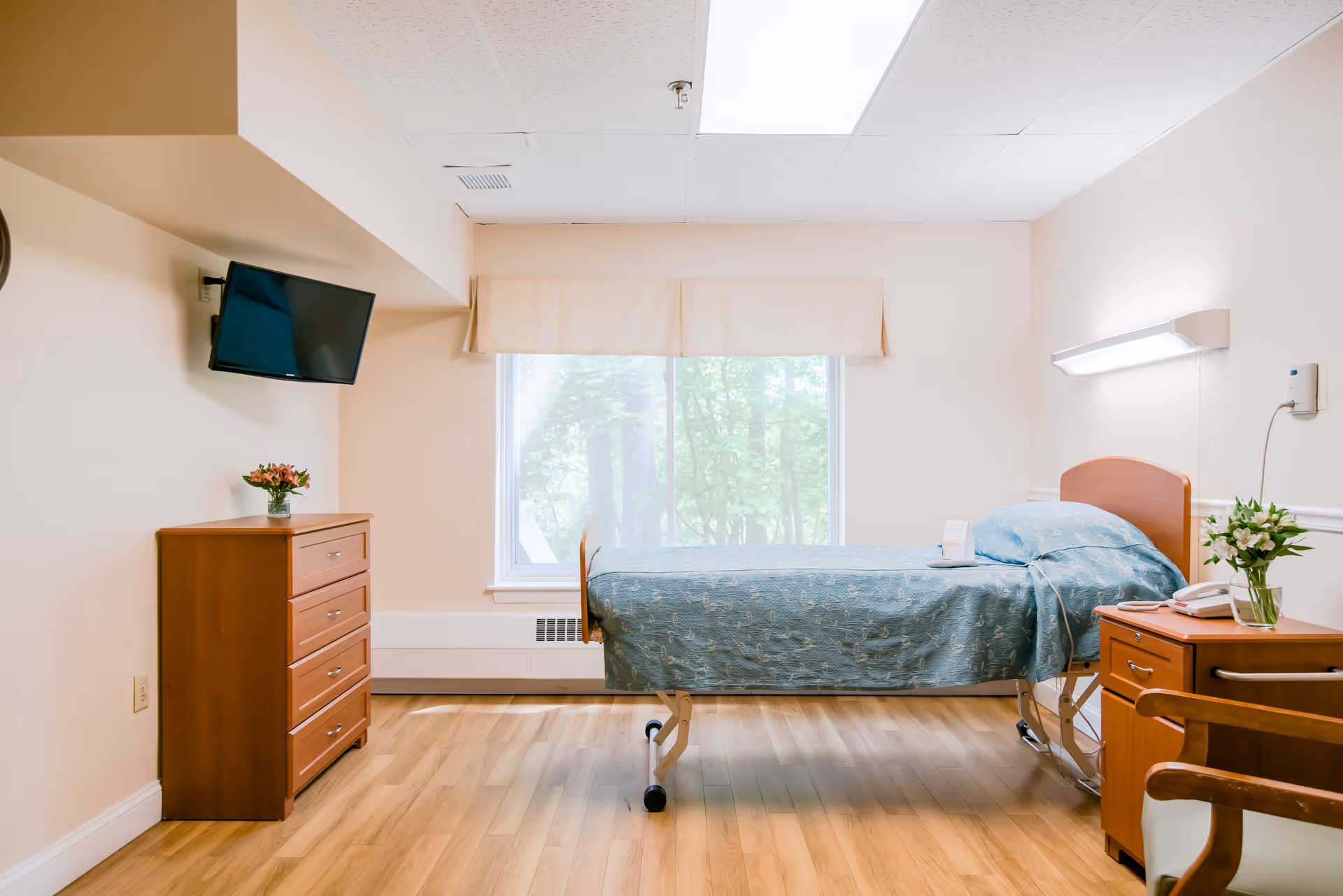 A clean and bright senior living bedroom with a single bed covered in a blue patterned bedspread. The room has light-colored walls and wood flooring. There is a wooden dresser with a small vase of flowers on top, a wall-mounted flat screen TV, a large window with a beige valance, and a wooden nightstand with a telephone and a vase of flowers. A wooden chair is partially visible in the foreground.