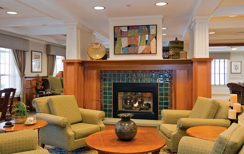 Cozy senior living common area with a fireplace surrounded by green tiles and wooden mantel. Four green upholstered armchairs are arranged around a wooden coffee table with a decorative vase. The room has large windows with curtains, framed artwork above the fireplace, and warm lighting.