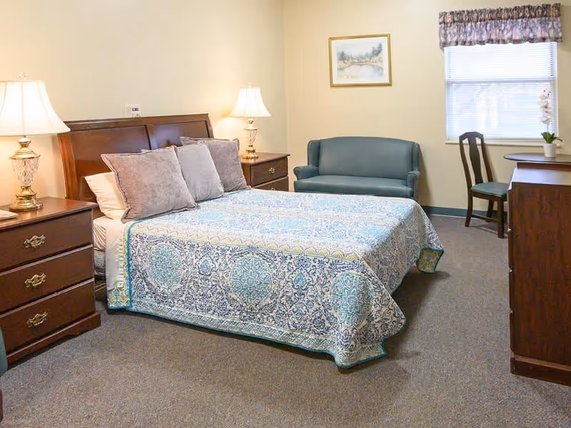 A neatly made bed with a patterned blue and white bedspread and three gray pillows in a senior living facility bedroom. The room features two wooden nightstands with lamps, a small blue loveseat, a wooden chair, a round table, a wooden dresser, a window with floral curtains, and a framed landscape painting on the wall.