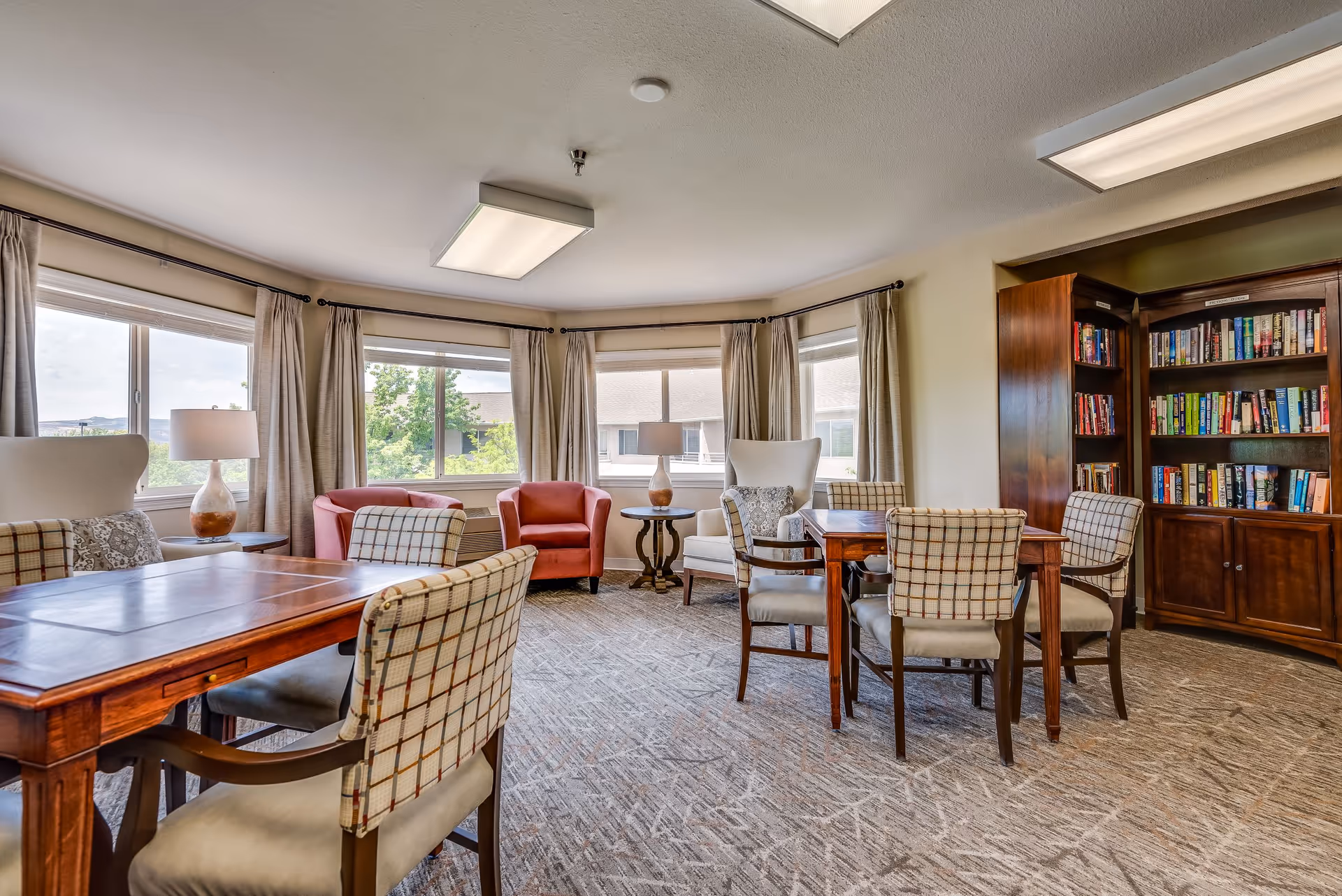 A cozy senior living common area with large windows letting in natural light. The room features two wooden tables surrounded by cushioned chairs with plaid upholstery. There are two red armchairs and two white armchairs near the windows, each accompanied by a side table with a lamp. A wooden bookshelf filled with books is positioned against the wall.