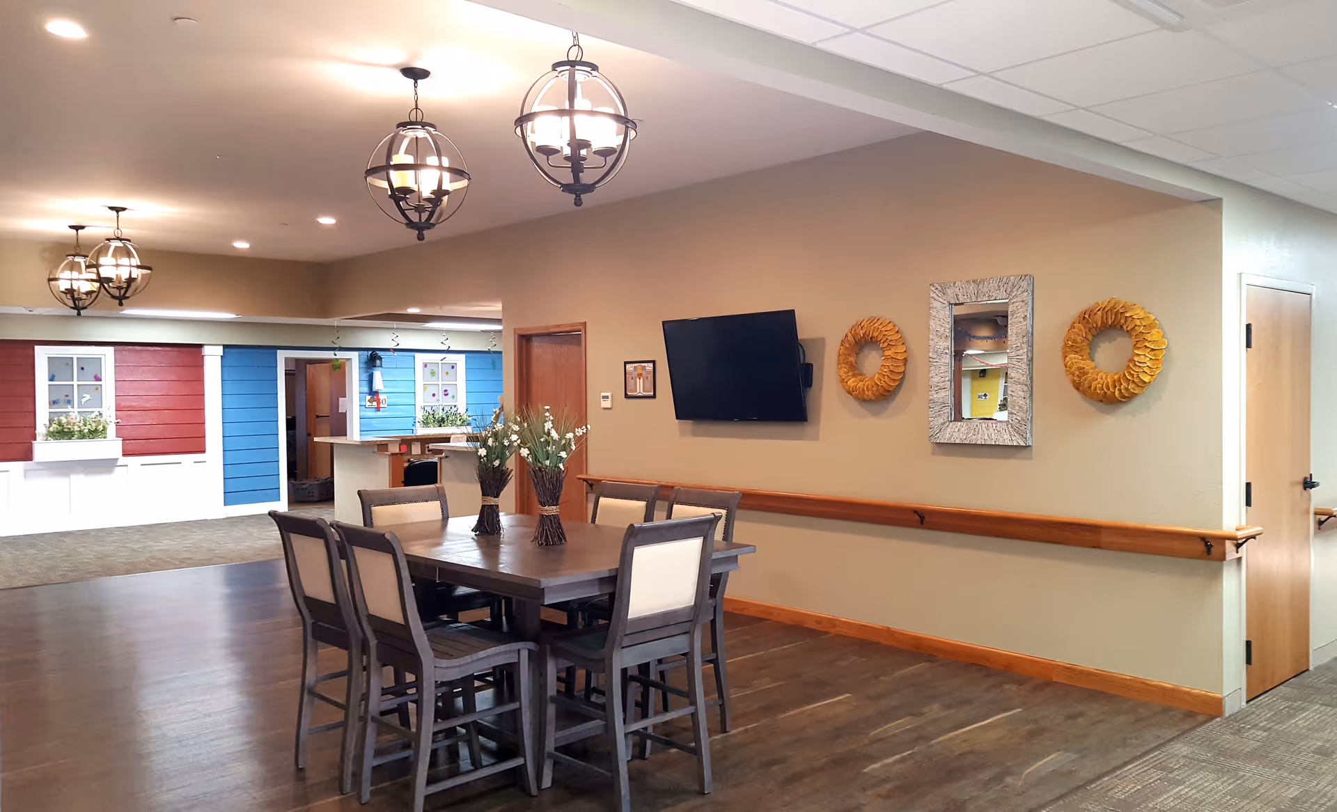 Interior view of a memory care facility common area with a wooden table and six chairs in the foreground. The walls are beige with a mounted flat-screen TV, two decorative wreaths, and a mirror. The floor is wood, and there are three hanging light fixtures on the ceiling. In the background, there are colorful red and blue wall panels with white window frames and flower boxes, and a reception desk area.