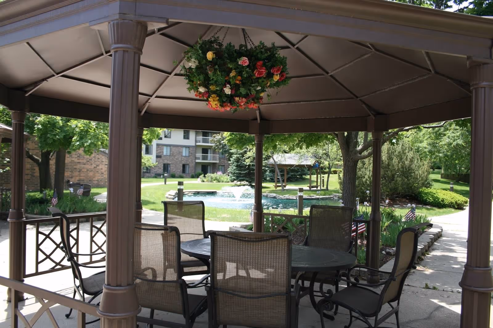 Outdoor gazebo with a round table and six chairs underneath, surrounded by greenery, trees, and a small pond with a fountain in the background. A hanging basket of colorful flowers is suspended from the gazebo ceiling.