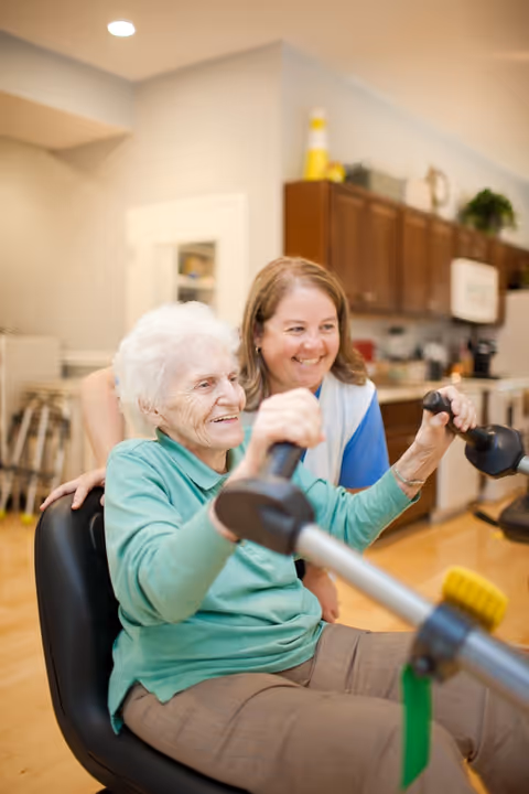 An elderly woman with white hair wearing a green shirt is seated and using exercise equipment with handles, smiling. A caregiver or staff member in a blue shirt and white vest is standing behind her, smiling and providing support. The background shows a kitchen area with wooden cabinets and various items on the counter.