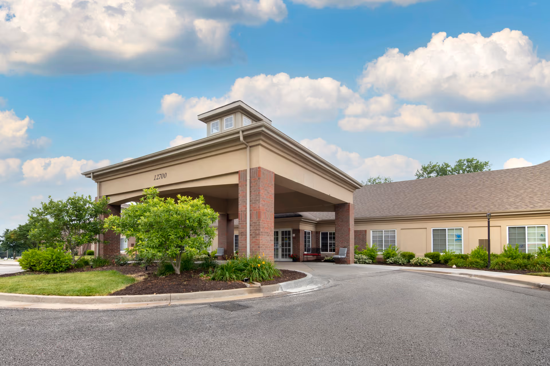 Front entrance canopy of a senior living facility with a drive-up circular driveway, landscaped beds and a partly cloudy sky.