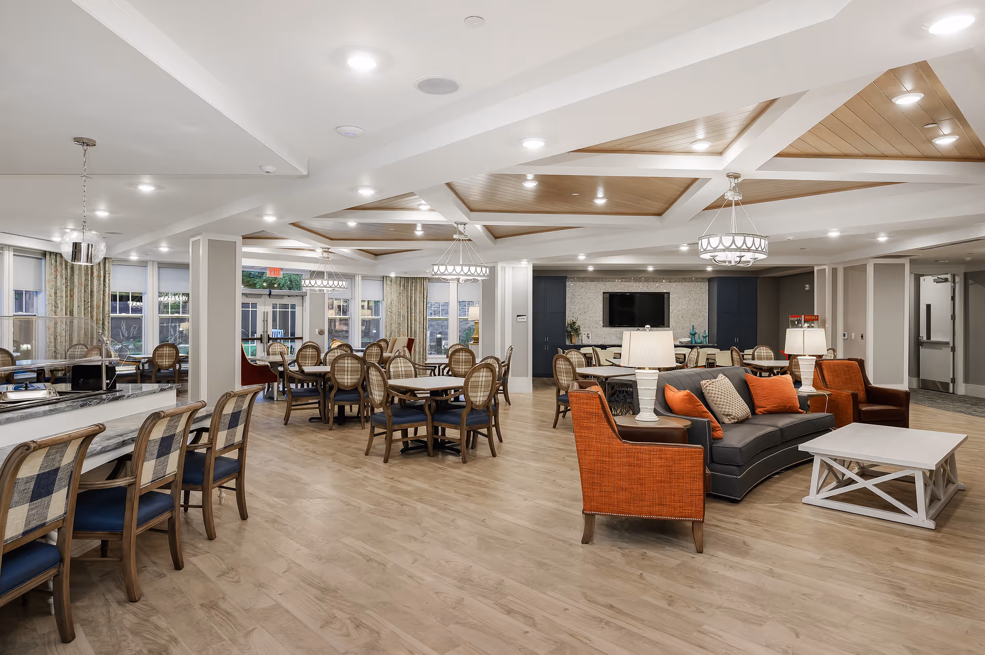 Spacious and well-lit senior living facility common area with multiple dining tables and chairs, a seating area with a dark sofa and orange armchairs, modern ceiling lights, and large windows with curtains.