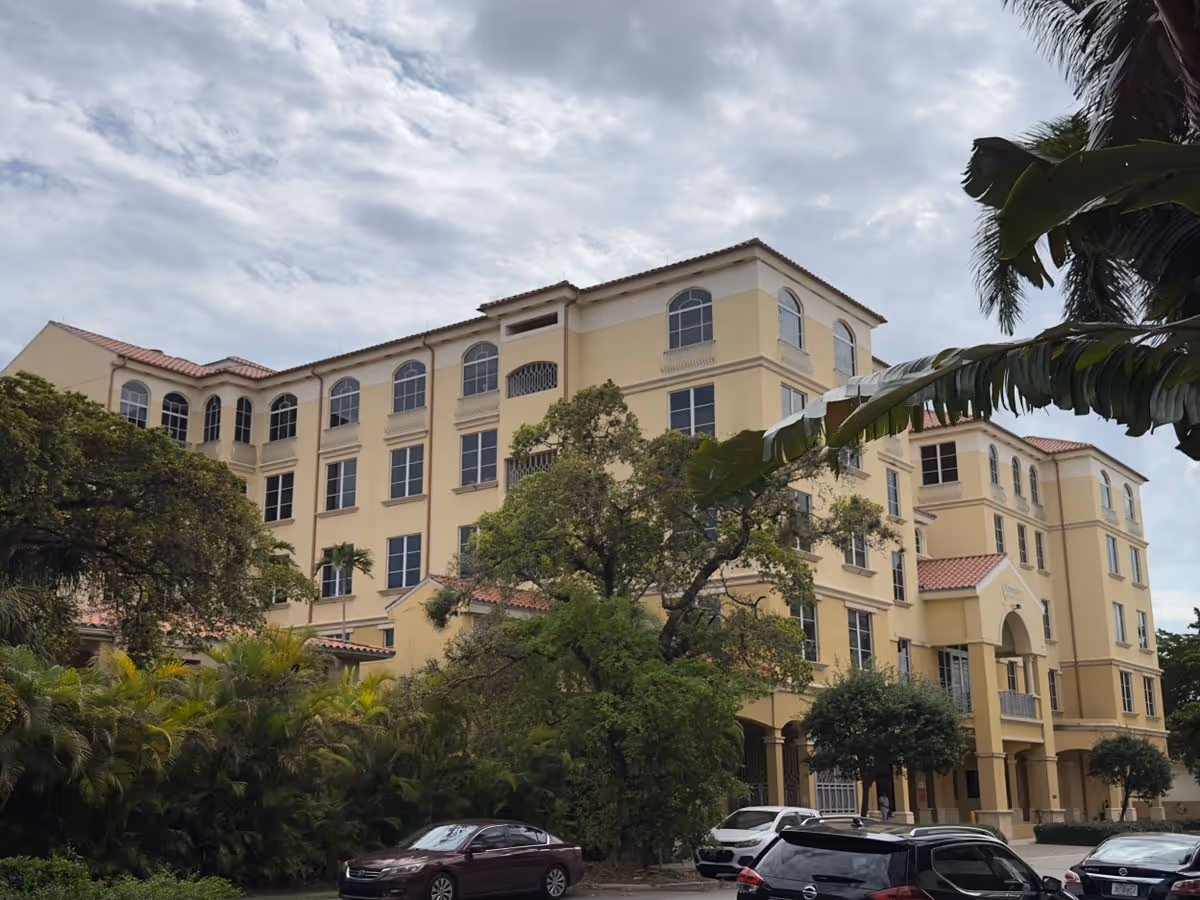 Front exterior of a multi-story Mediterranean-style yellow building with arched windows, landscaping, and parked cars in front.