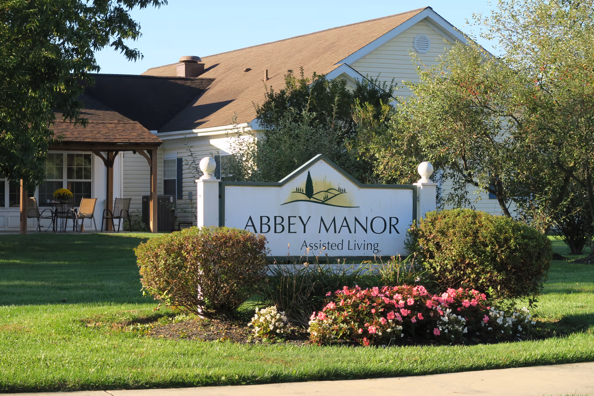 Exterior view of Abbey Manor Assisted Living facility with a white sign displaying the facility name surrounded by bushes and flowers. The building is light-colored with a brown roof, and there is a covered patio area with chairs and a table visible on the left side.