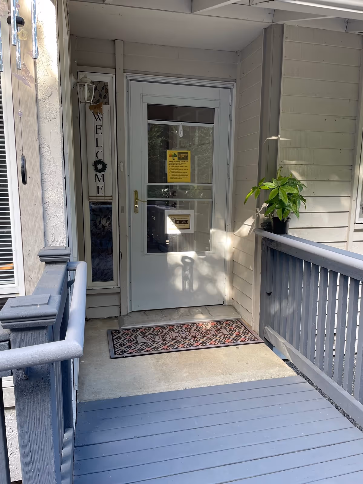 Entrance to a facility with a white door and a glass storm door. There is a vertical welcome sign on the left side of the door and a welcome doormat on the concrete porch. A small potted plant is placed on the right railing of the porch. The porch has gray wooden railings and floorboards.