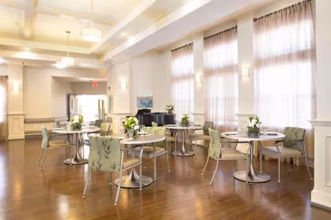 Sunlit senior living dining room with round tables, chairs, and floral centerpieces.