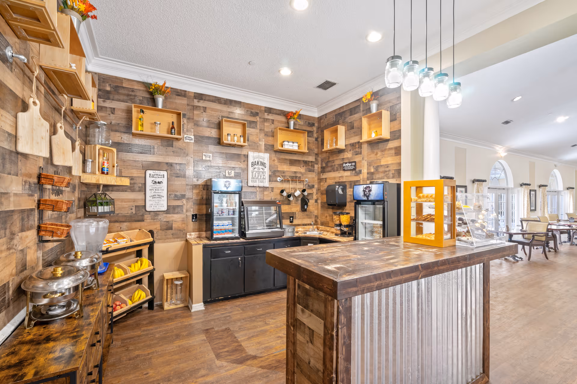 A rustic-style kitchen area with wooden walls and shelves, featuring a countertop with hanging mason jar lights, a display case with pastries, a beverage cooler, a coffee machine, and various kitchen utensils. The space opens into a dining area with tables and chairs near large arched windows.