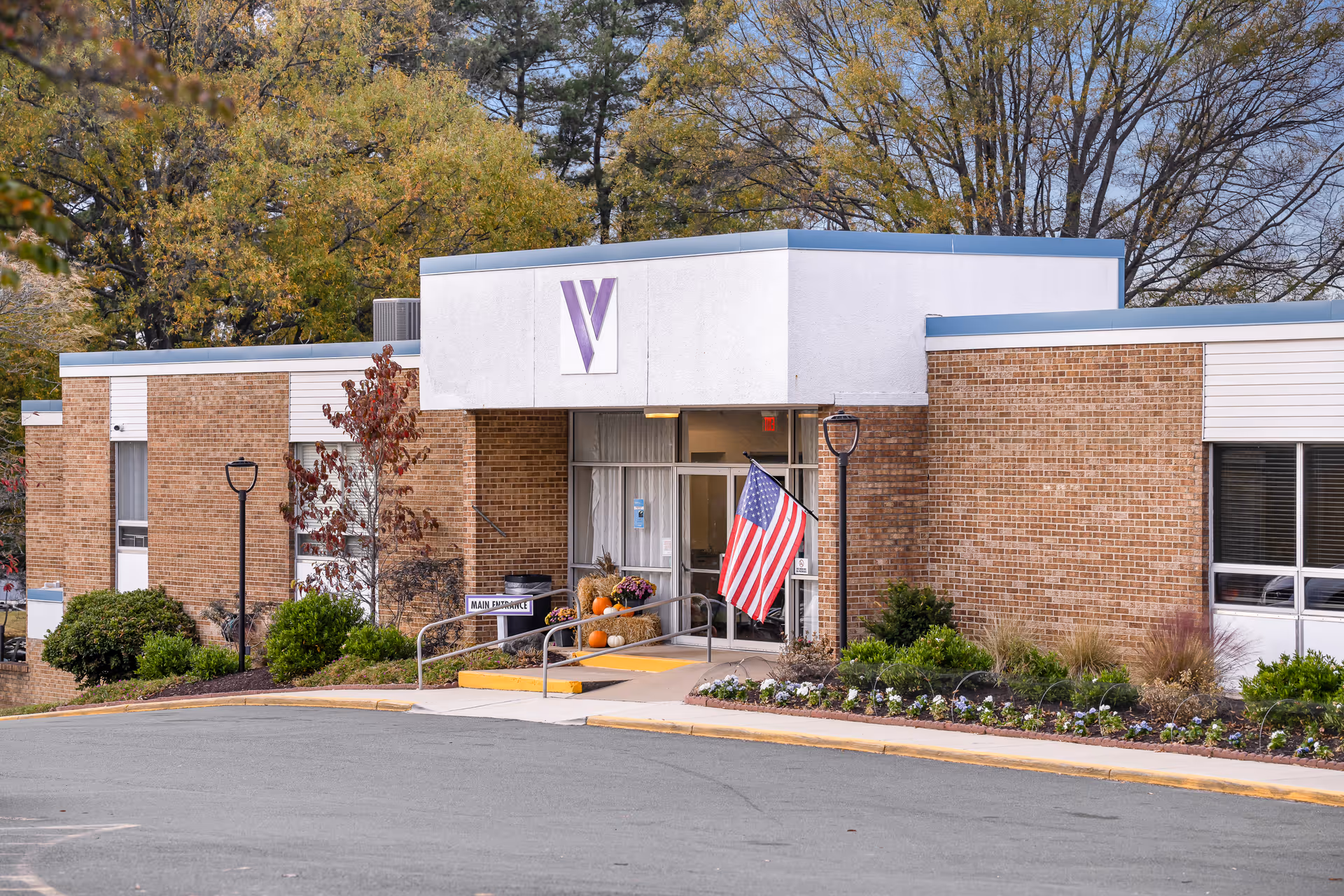 Front entrance of a brick senior living building with an American flag, wheelchair ramp, fall decorations, and trees in the background.