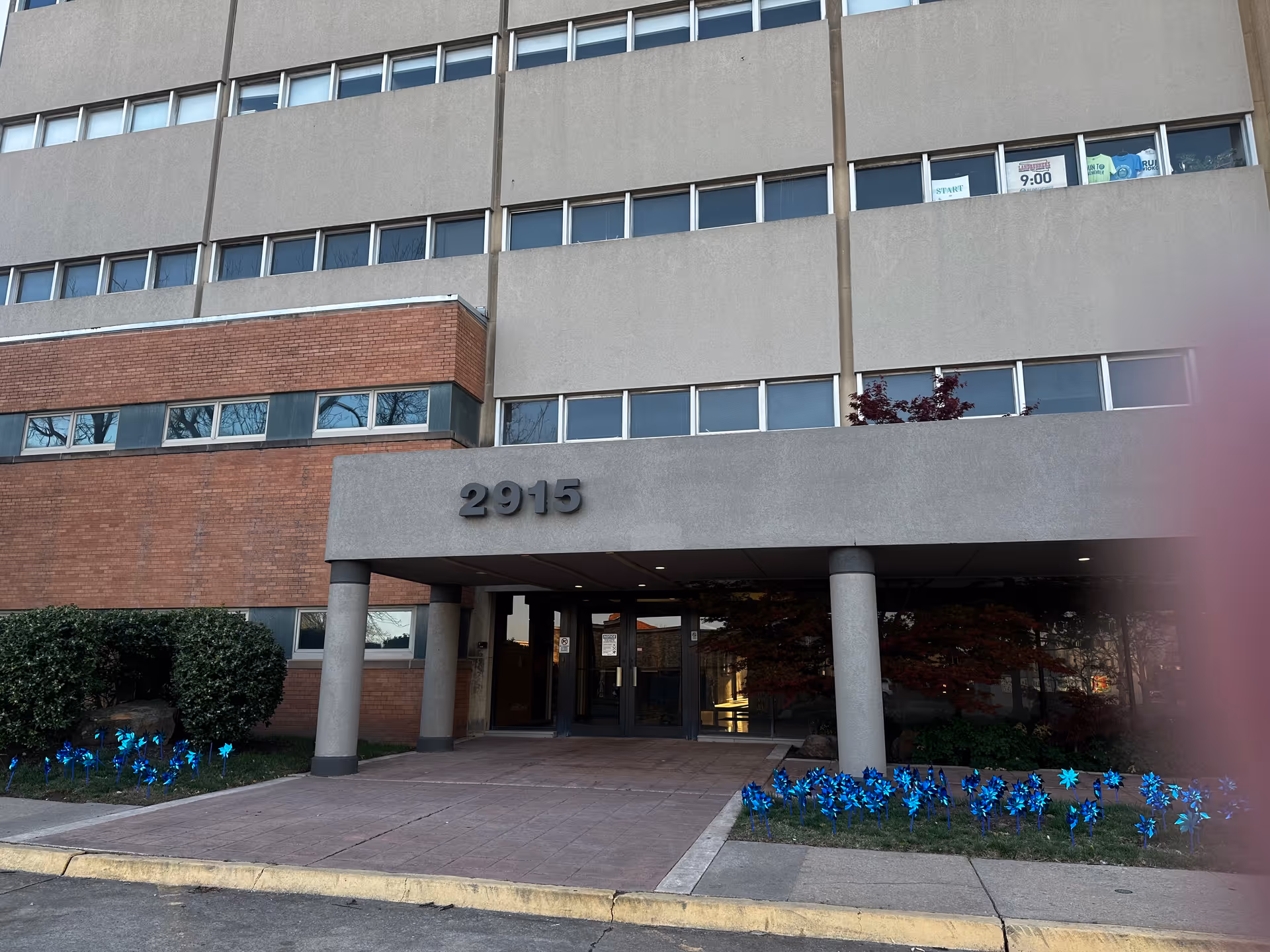 Exterior front entrance of a multi-story building with the number 2915 displayed above the entrance. The building has a combination of brick and concrete facade with several windows. There are blue pinwheels planted in the flower beds on either side of the entrance and some bushes. A partial finger is visible on the right side of the image.