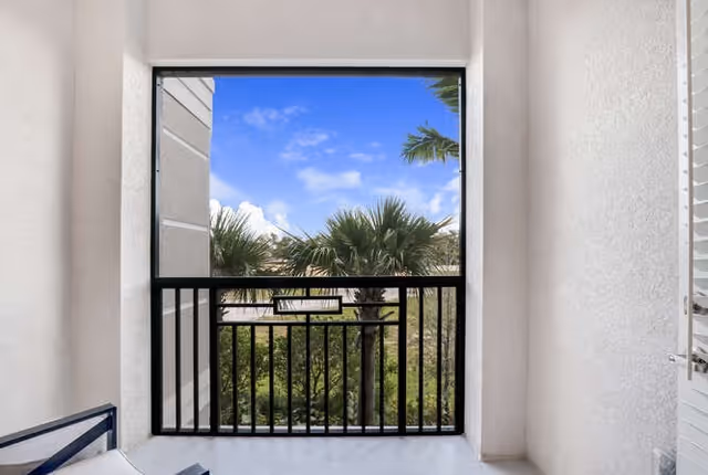 Covered balcony with a black metal railing overlooking palm trees and a bright blue sky.