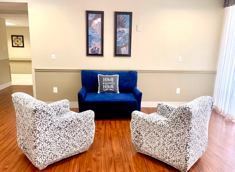Small seating area with a blue loveseat, two patterned armchairs on a wood floor and framed artwork on the wall.