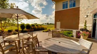 Outdoor patio area at Tallgrass Creek Senior Living Community with round wooden tables and chairs under large umbrellas, surrounded by plants and greenery, with a building wall on one side and a scenic view of a grassy field and partly cloudy sky in the background.