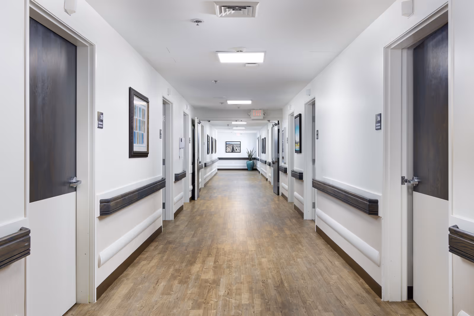 A long, clean hallway in a senior living facility with wooden floors, white walls, and multiple closed doors on both sides. The hallway is well-lit with ceiling lights and has handrails along the walls. There are framed pictures hanging on the walls and a potted plant at the far end of the corridor.