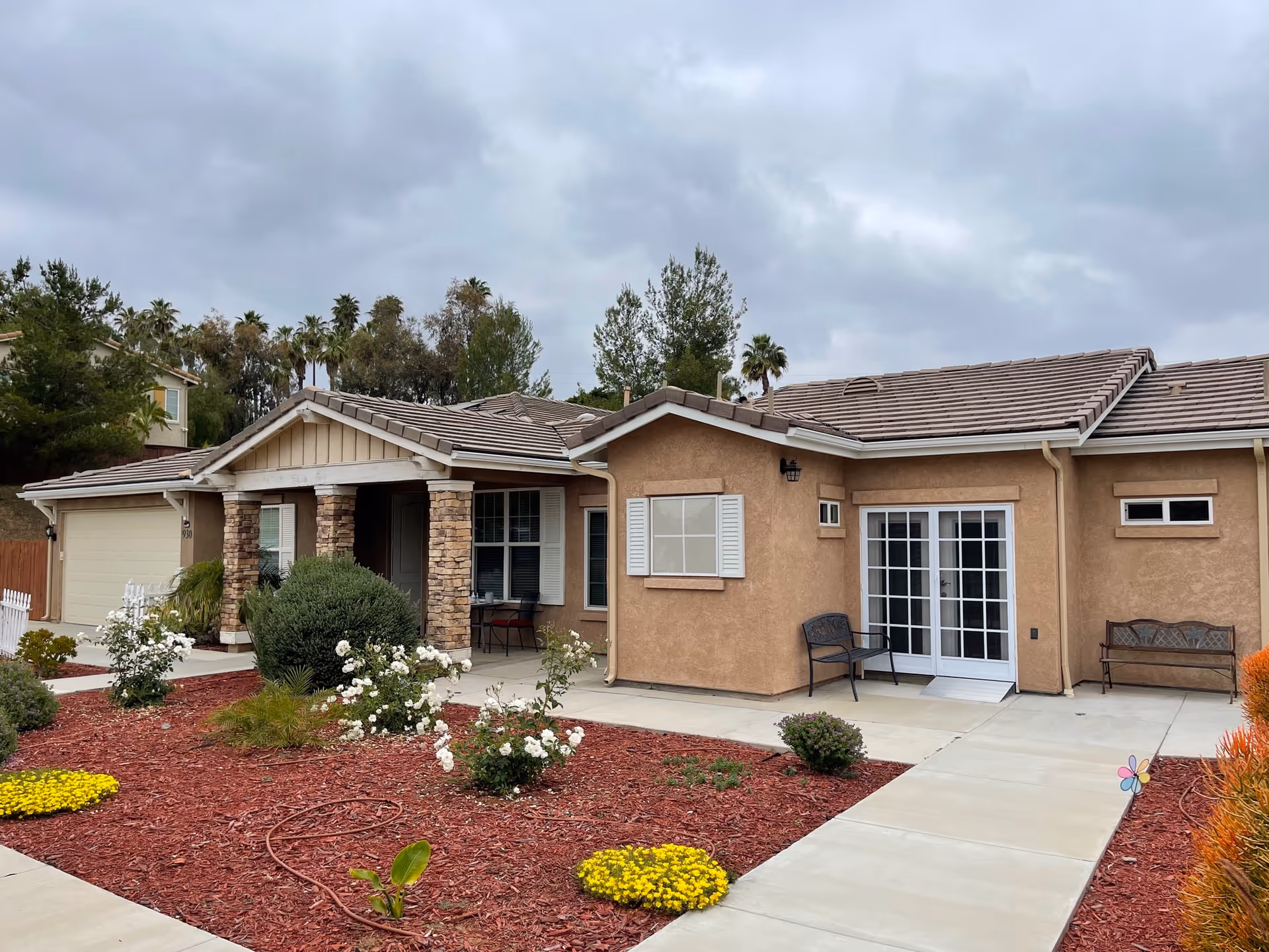 Exterior view of a single-story residential building with a beige stucco finish and a tiled roof. The front yard features a landscaped garden with red mulch, white and yellow flowers, and shrubs. There is a concrete pathway leading to the entrance, which has stone pillars and a covered porch. Two benches are placed near the entrance and a set of white-framed glass doors. The sky is overcast with gray clouds.