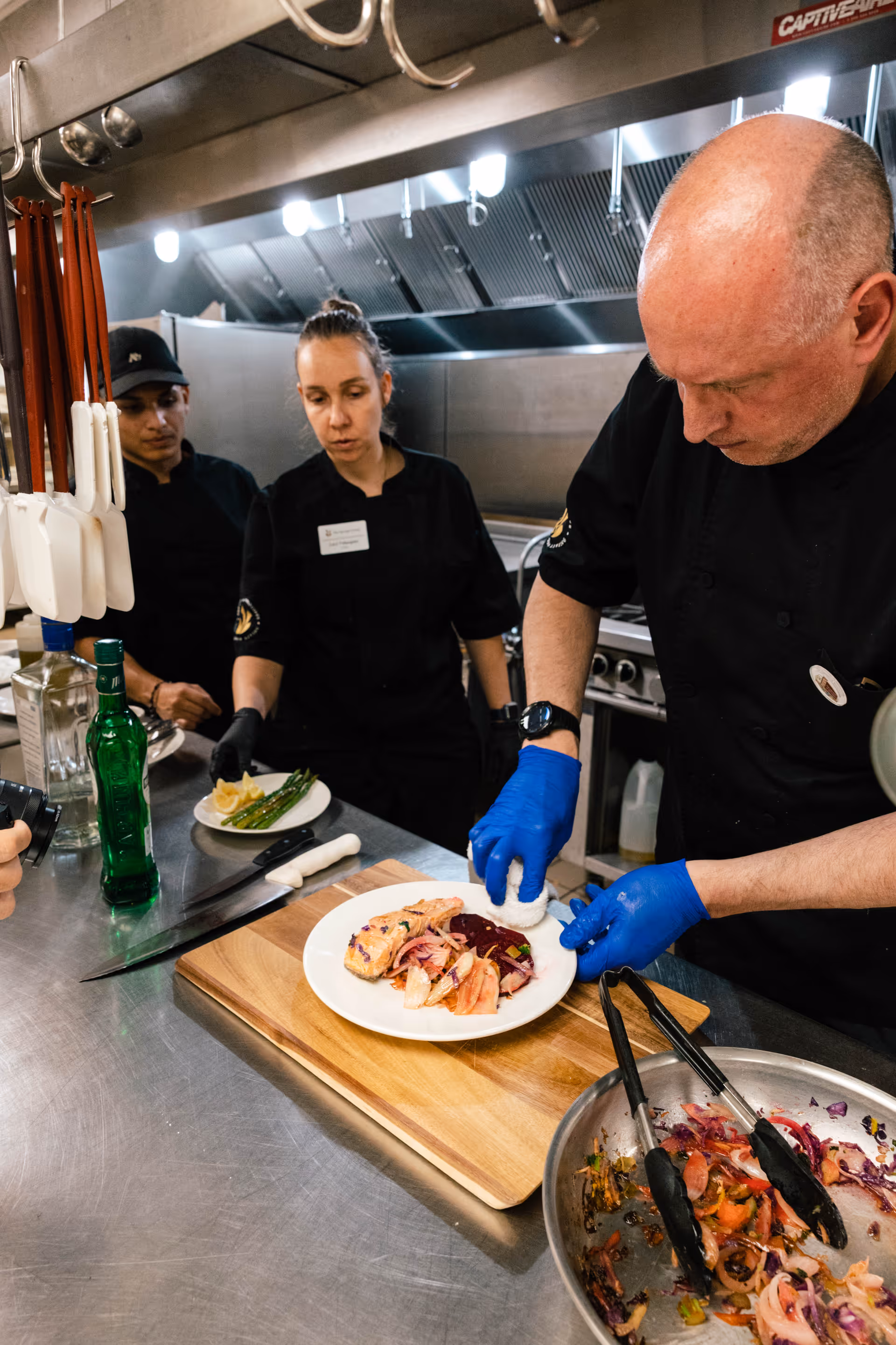 Three chefs in a commercial kitchen preparing and plating food. One chef is placing a white food item on a plate with cooked salmon and vegetables, while the other two chefs observe. Various kitchen utensils and ingredients are visible on the stainless steel counter.