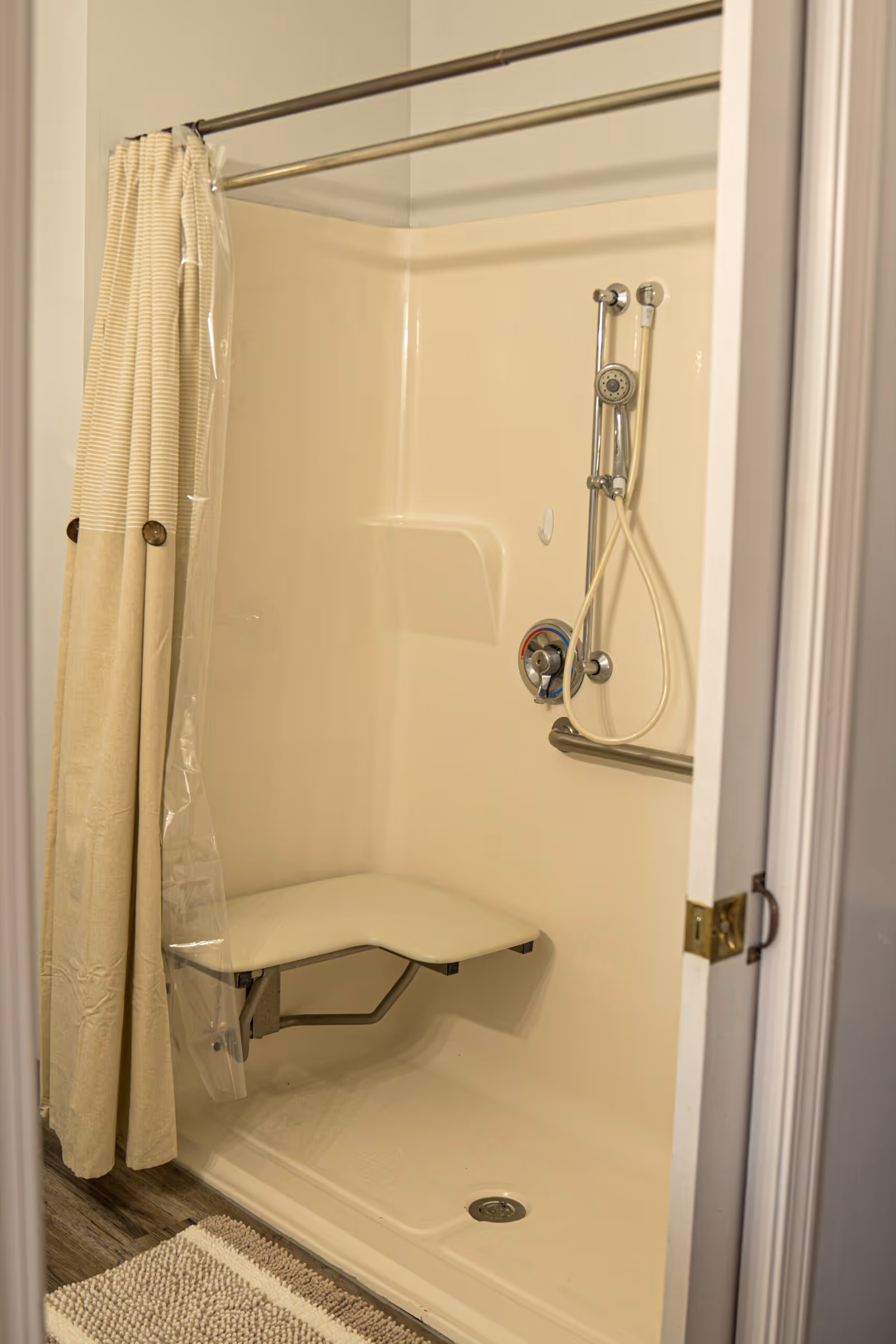 A beige accessible shower stall with a fold-down seat, a handheld showerhead attached to a sliding bar, and a grab bar. The shower has a beige curtain with a clear plastic liner and a textured bath mat on the floor outside the shower.