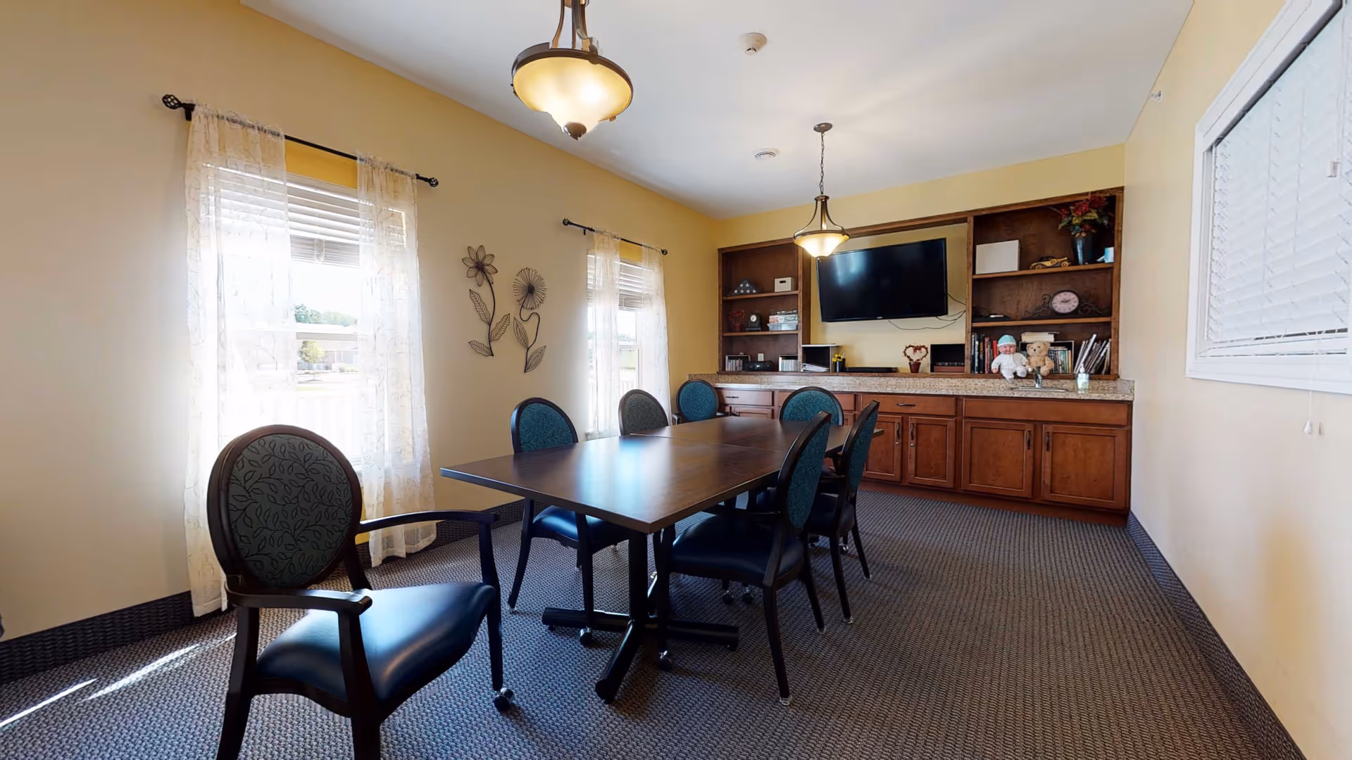 A well-lit room with a rectangular wooden table surrounded by six padded chairs with armrests. The room has two windows with sheer white curtains letting in natural light. On the far wall, there is a built-in wooden cabinet with shelves, decorative items, and a mounted flat-screen TV. The walls are painted a soft yellow, and the floor is carpeted in a dark pattern. Two ceiling light fixtures hang above the table.