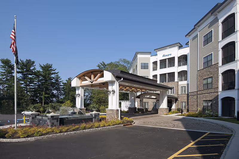 Exterior view of Allegro Senior Living facility in Harrington Park, NJ, showing a covered entrance with white pillars and wooden beams, a flagpole with an American flag, landscaped garden beds, and a multi-story building with balconies and stone accents under a clear blue sky.
