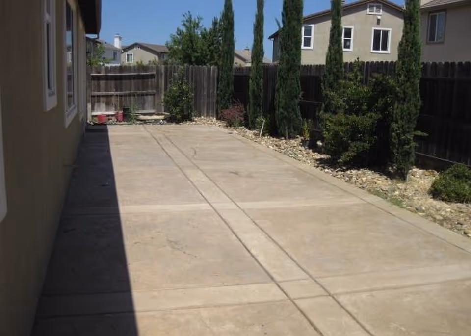 Concrete side yard or driveway beside a house with a wooden fence, tall narrow trees, and neighboring houses visible in the background.