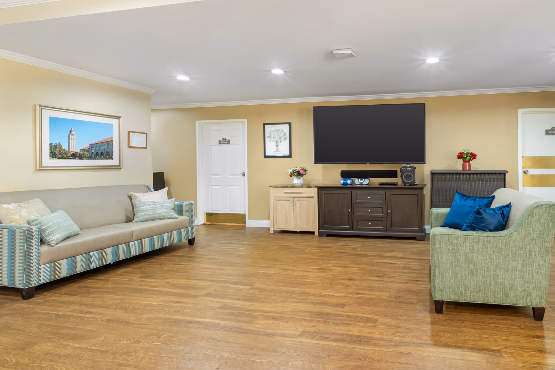 Spacious senior living lounge with sofas, a wall-mounted TV above a media console, decorative pillows and flowers, and hardwood floors.