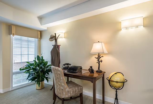 A cozy corner office space with a wooden desk and a cushioned chair. On the desk are a vintage typewriter, an old-fashioned rotary phone, a small dog figurine, and a lamp with a tree-branch styled base. To the right of the desk is a globe on a stand. A coat rack with a hat and coat is positioned near a large window with blinds, and a green potted plant sits beside the window. The walls are painted light beige and two wall-mounted lights illuminate the area.