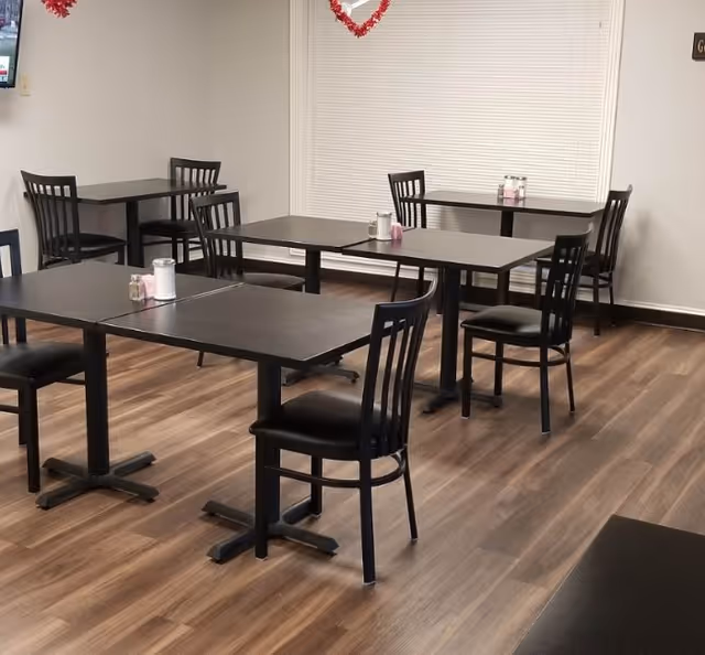Empty dining area with several black tables and chairs on wood-look flooring and condiments on each table.