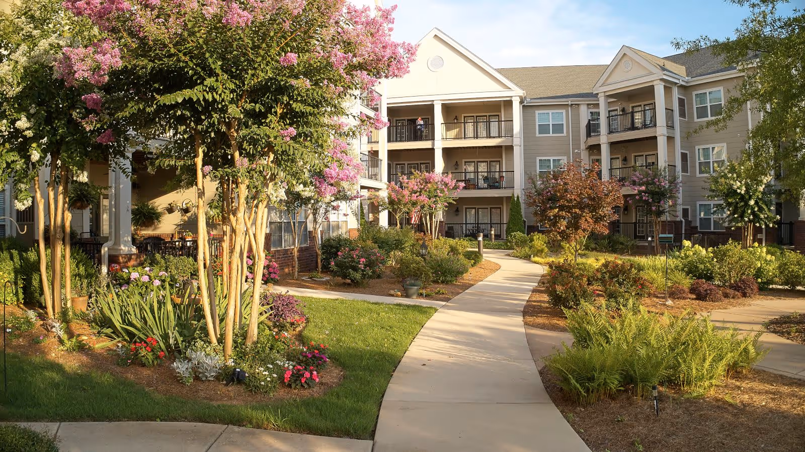 A landscaped outdoor courtyard area with a paved walkway winding through green grass, flowering plants, and trees with pink and white blossoms. Surrounding the courtyard are multi-story residential buildings with balconies and large windows under a clear blue sky.