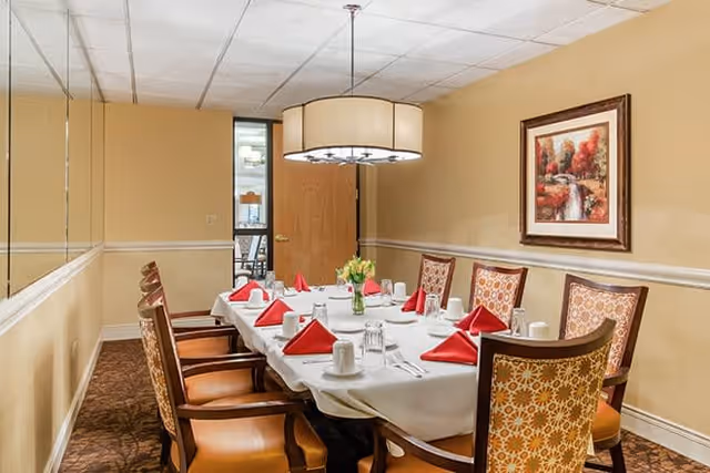 A dining room with a rectangular table set for eight people. The table is covered with a white tablecloth and has red folded napkins, white cups, and silverware. There is a vase with yellow flowers in the center. The room has beige walls, a large circular ceiling light, and a framed painting of a waterfall and autumn trees on the wall.