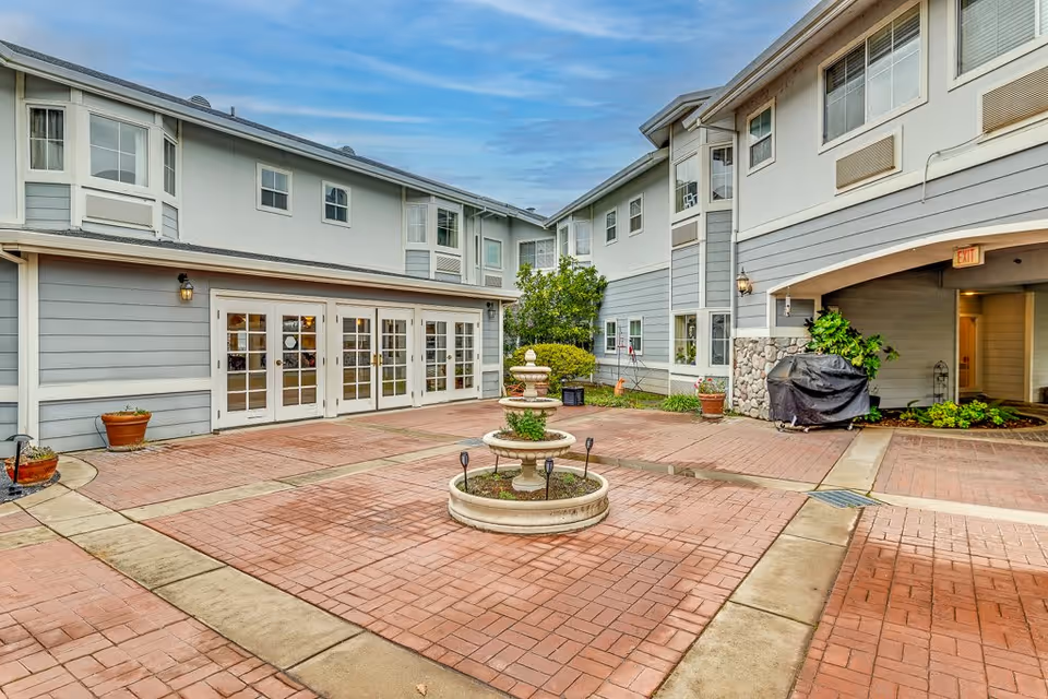 Outdoor courtyard area of a senior living facility with a central two-tiered fountain surrounded by brick paving. The courtyard is flanked by two light gray buildings with multiple windows and white trim. There are potted plants and greenery around the edges, and a covered walkway on the right side. The sky is partly cloudy.