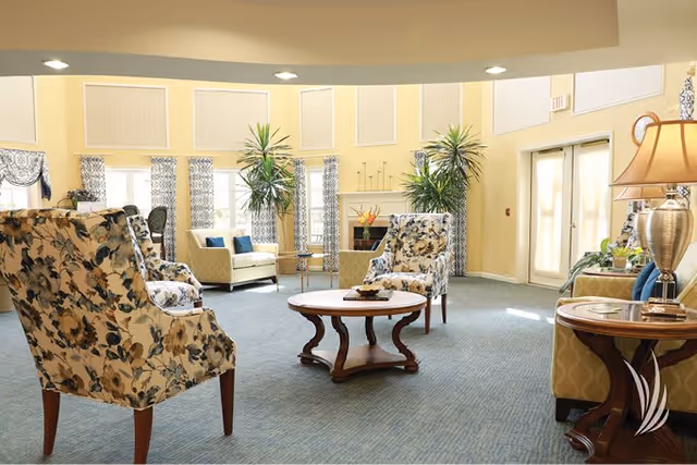 A bright and spacious senior living community lounge with floral patterned armchairs, a round wooden coffee table, large windows with patterned curtains, tall green plants, and soft yellow walls. The room is well-lit with recessed ceiling lights and a table lamp on a side table.