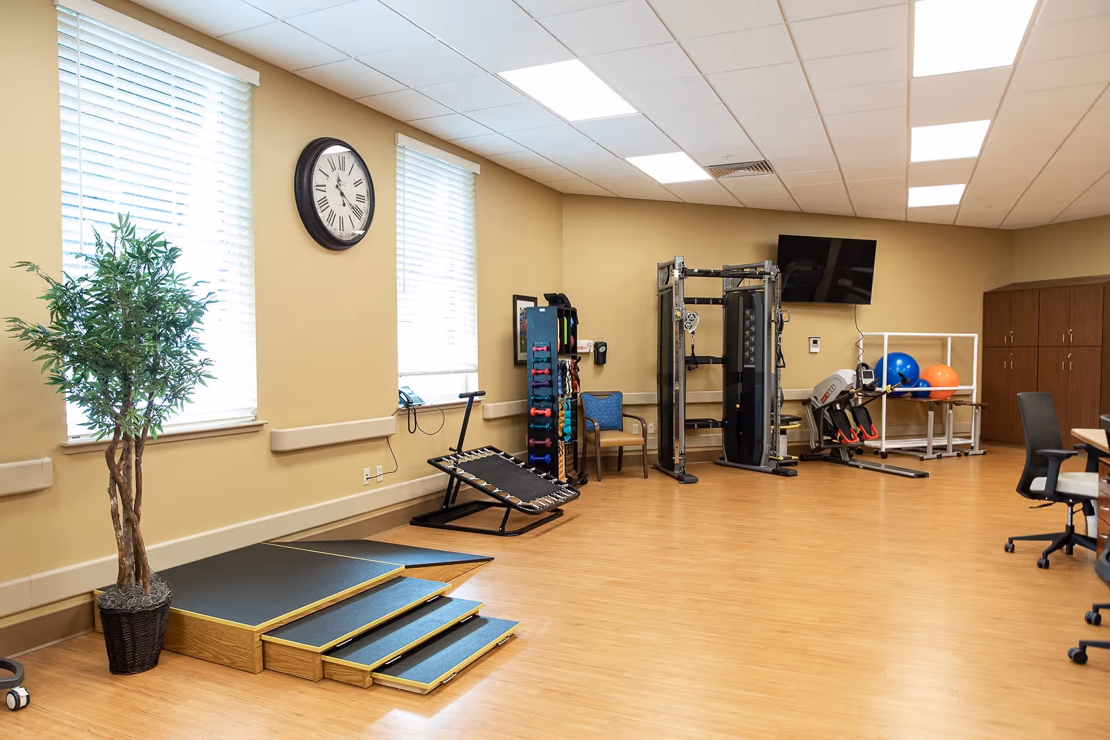 A spacious rehabilitation or physical therapy room with wooden flooring, exercise equipment including a multi-gym machine, resistance bands, exercise balls, a small trampoline, and a ramp with steps. There are two large windows with blinds, a wall clock, a potted plant, a chair, and a wall-mounted TV. The room has beige walls and a drop ceiling with fluorescent lighting.