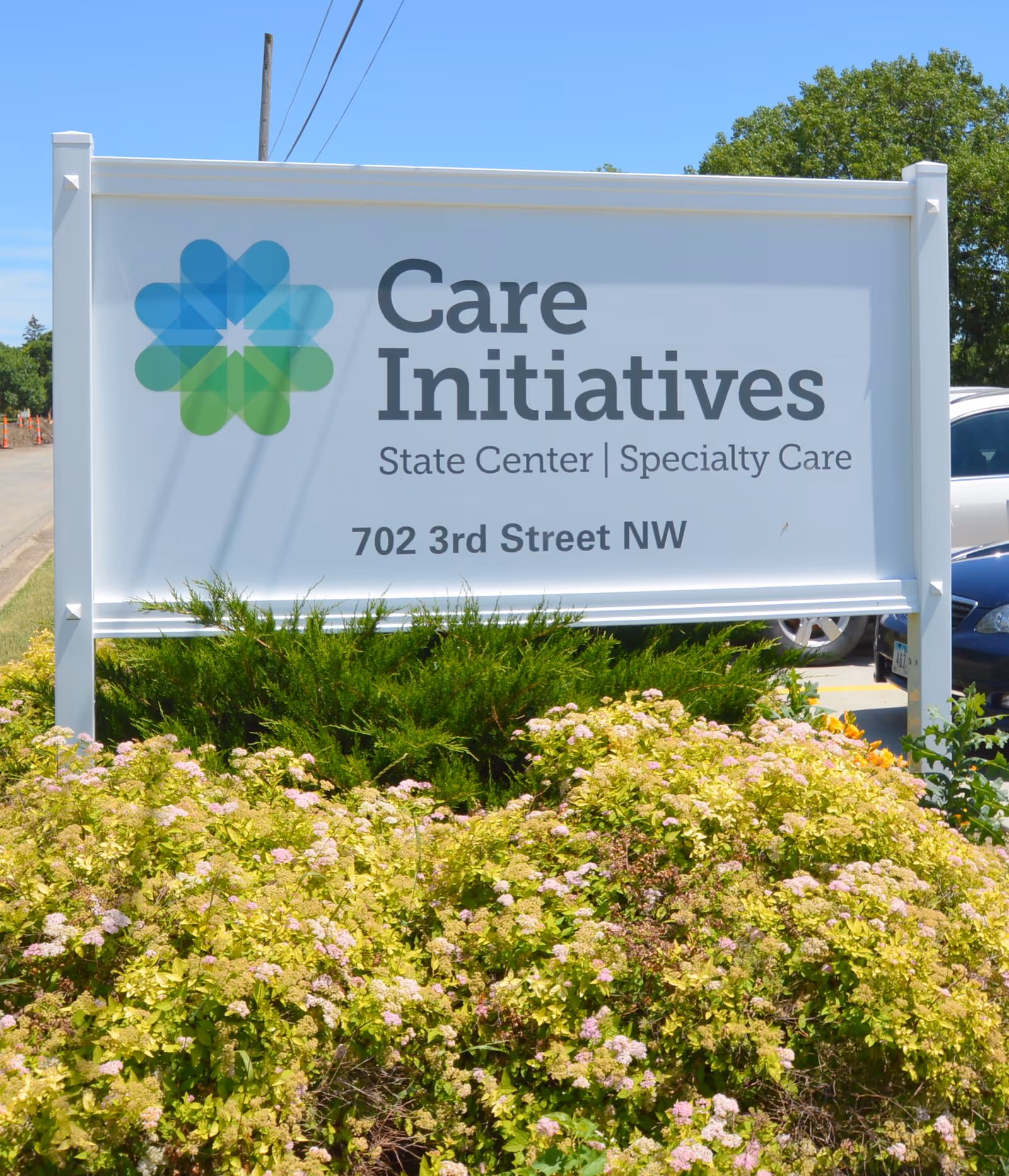 Outdoor sign for Care Initiatives State Center Specialty Care located at 702 3rd Street NW, surrounded by green bushes and flowering plants with a clear blue sky in the background.