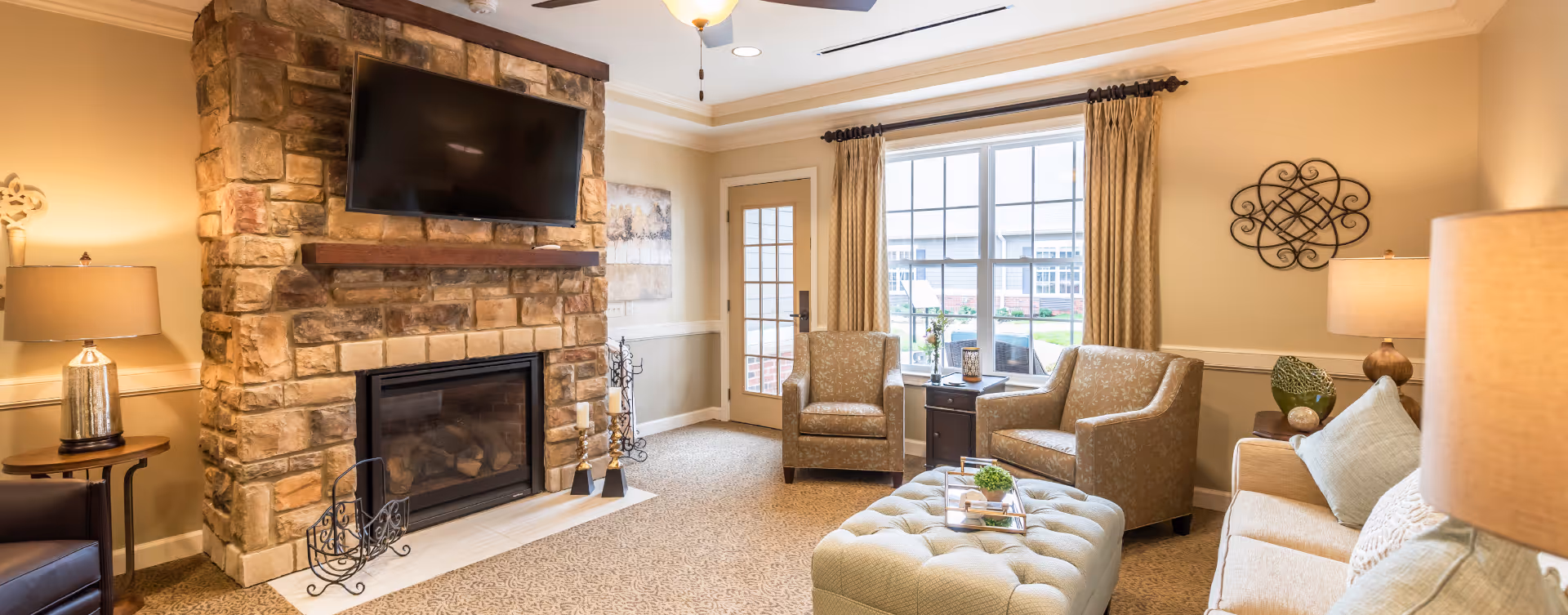 A cozy living room with a stone fireplace and a mounted flat-screen TV above it. The room features two patterned armchairs, a beige sofa with cushions, a tufted ottoman with a tray holding decorative items, and two table lamps on either side. A large window with curtains and a glass door provide natural light, and decorative wall art is visible.