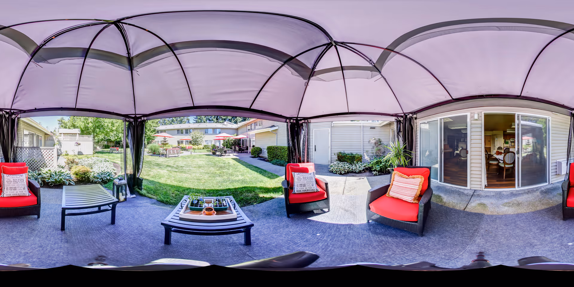 Covered outdoor patio with red cushioned chairs and coffee tables overlooking a grassy courtyard and sliding glass doors to the building.