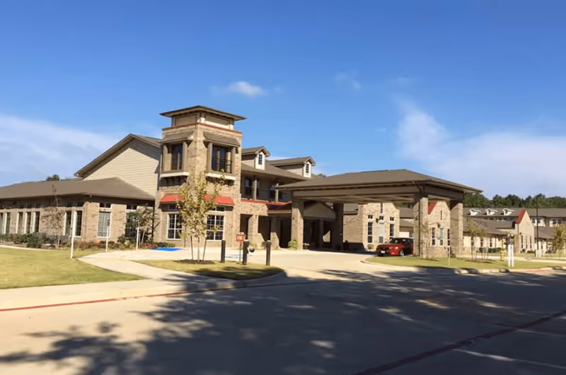 Exterior view of a senior living facility building with stone and beige siding, a covered entrance, and a driveway. The sky is clear and blue with some clouds, and there is a grassy area with small trees in front of the building.