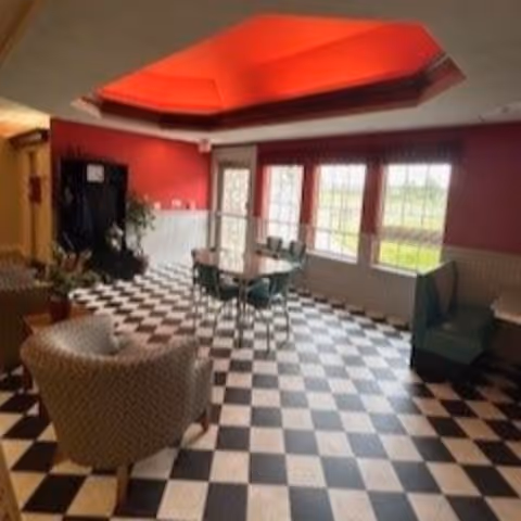Interior room with black and white checkered floor, red walls, and a red recessed ceiling. The room contains a round table with four chairs, two patterned armchairs, a green booth seat, and large windows letting in natural light.